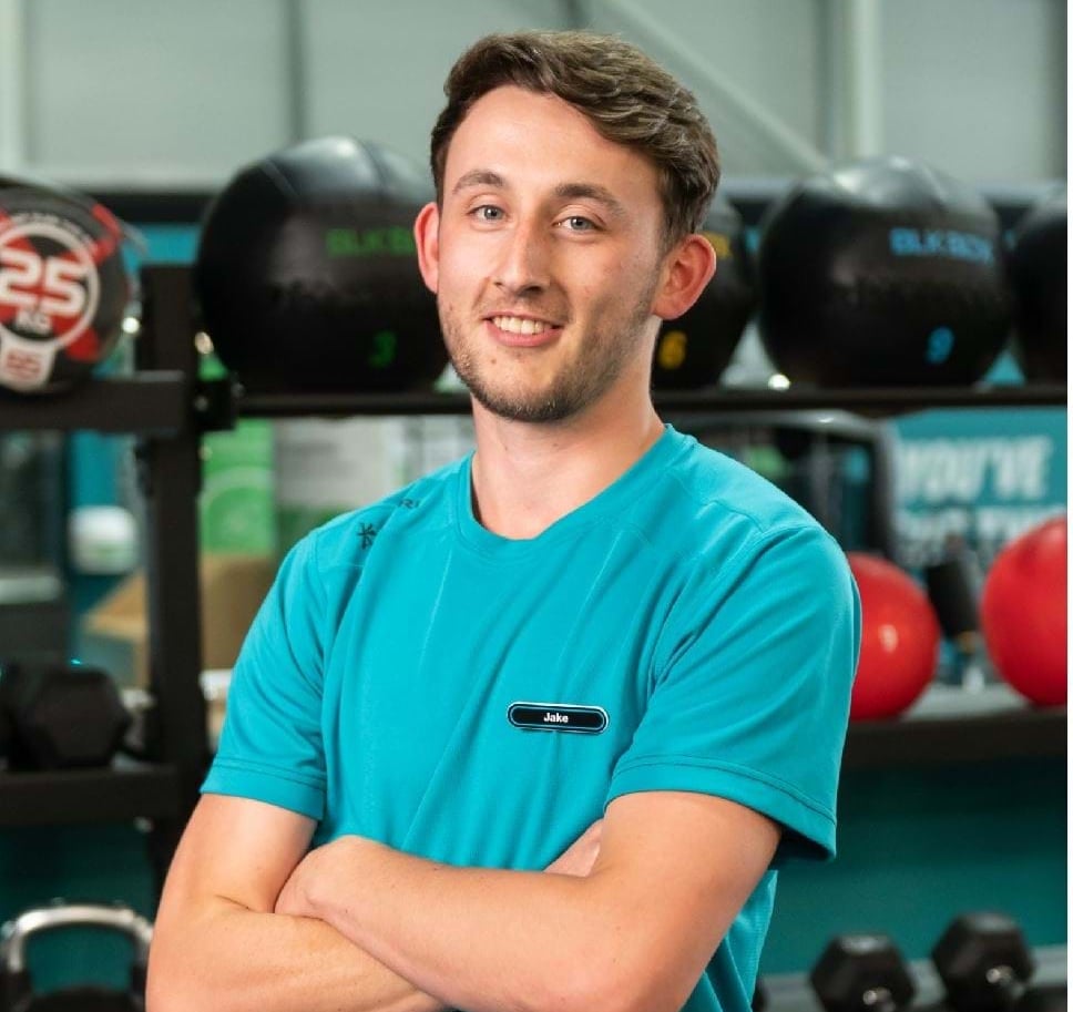 A male gym staff member named Jake smiles and crosses his arms in front of gym equipment, including medicine balls and dumbbells.