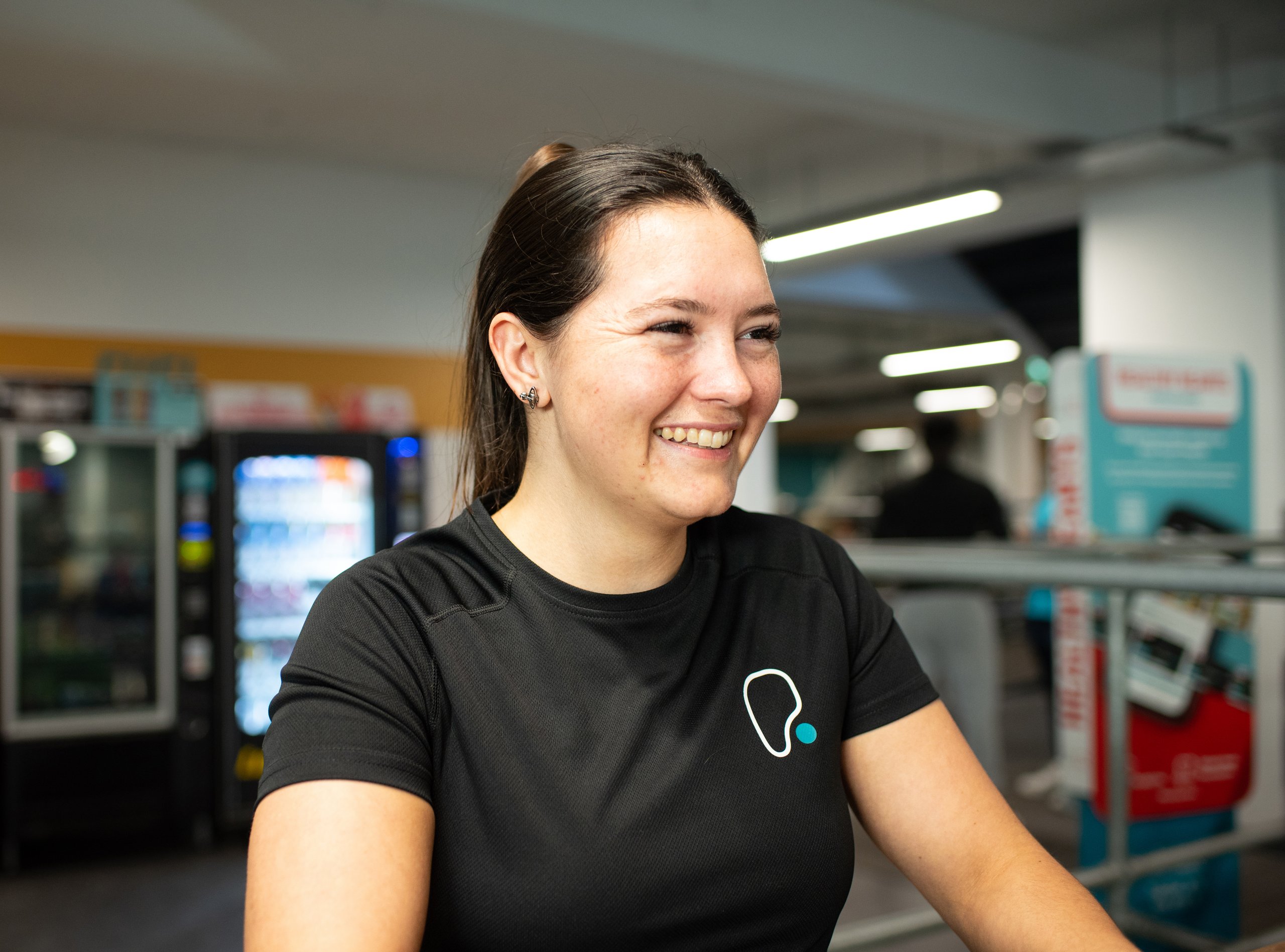 A woman with dark hair pulled back in a ponytail smiles, wearing a black t-shirt with a white and blue logo. She is in a gym setting with a vending machine and fitness equipment blurred in the background.
