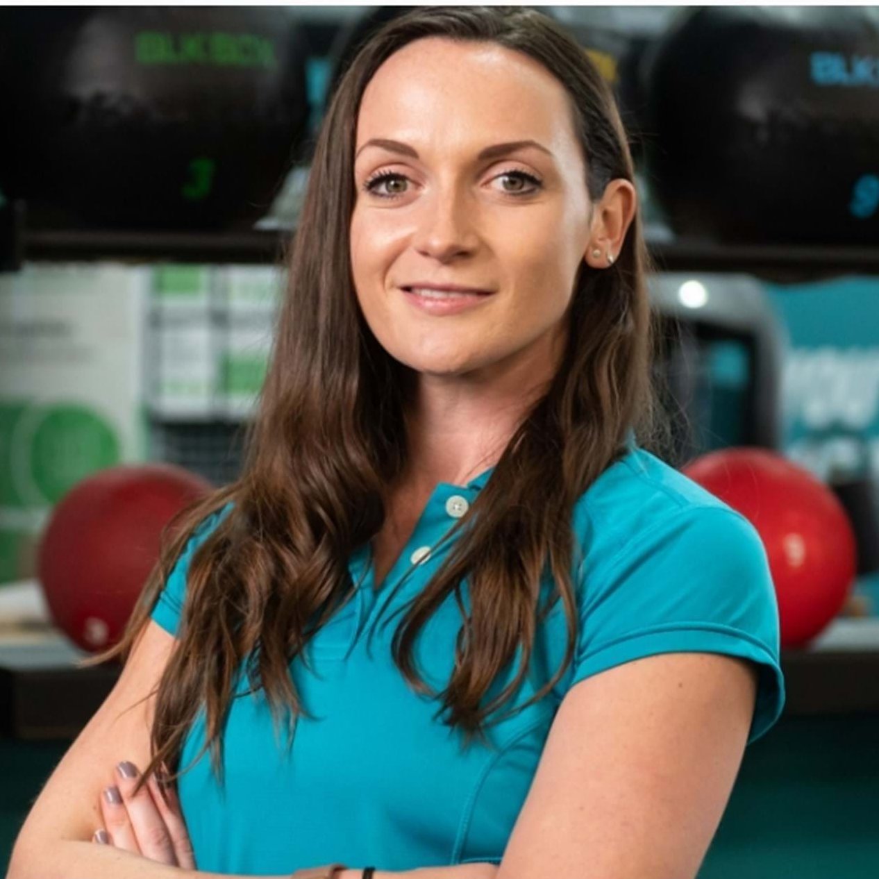 A female gym manager with long brown hair, wearing a teal polo shirt, standing in a gym.
