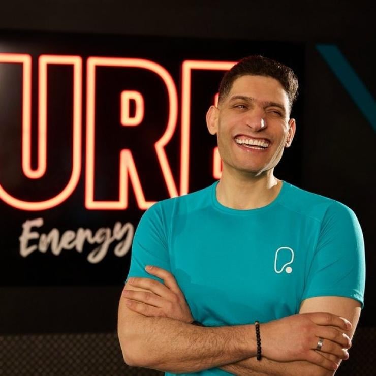 A smiling man in a teal athletic shirt with arms crossed in front of a glowing neon sign.