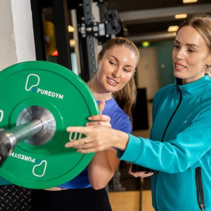A female gym instructor in a blue jacket guiding a client on using a PureGym weight plate.