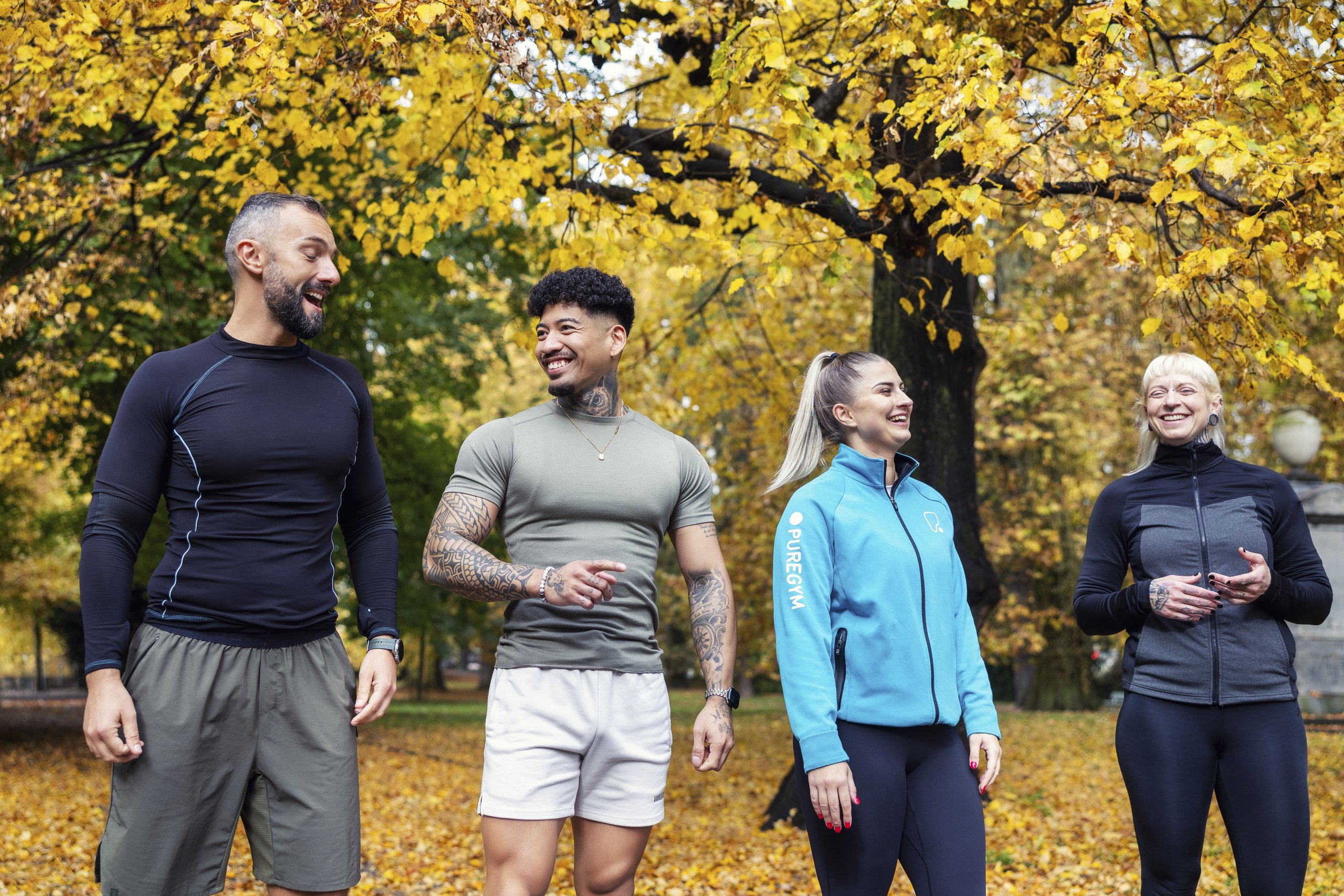 Four diverse fitness trainers talking and laughing outdoors against a backdrop of golden autumn trees.