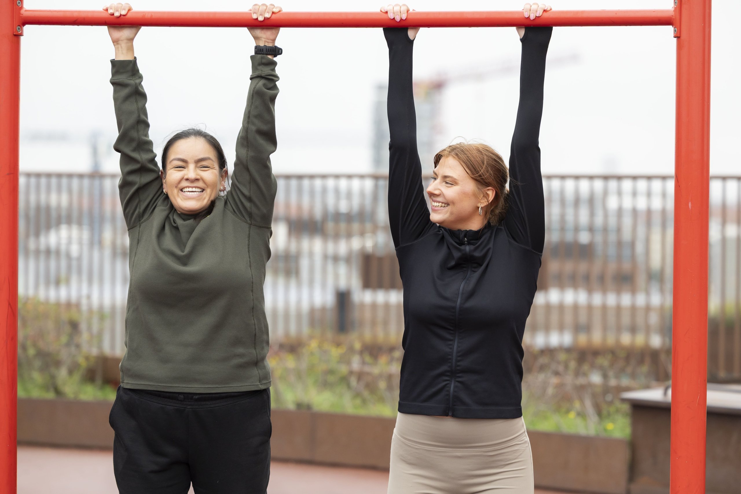 Two smiling women hanging from a red outdoor pull-up bar during a workout.