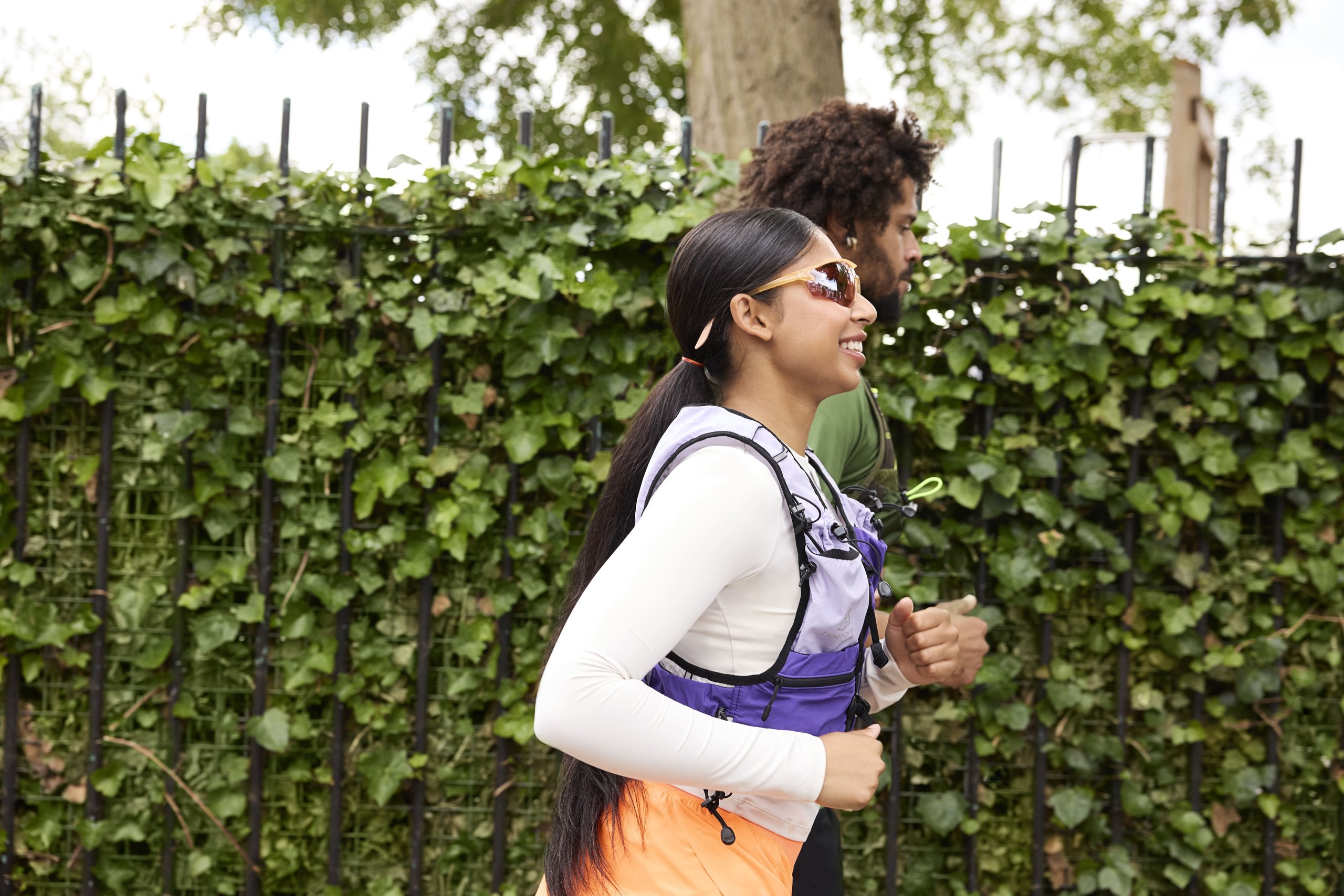 A woman and a man running together past a metal fence covered in ivy.