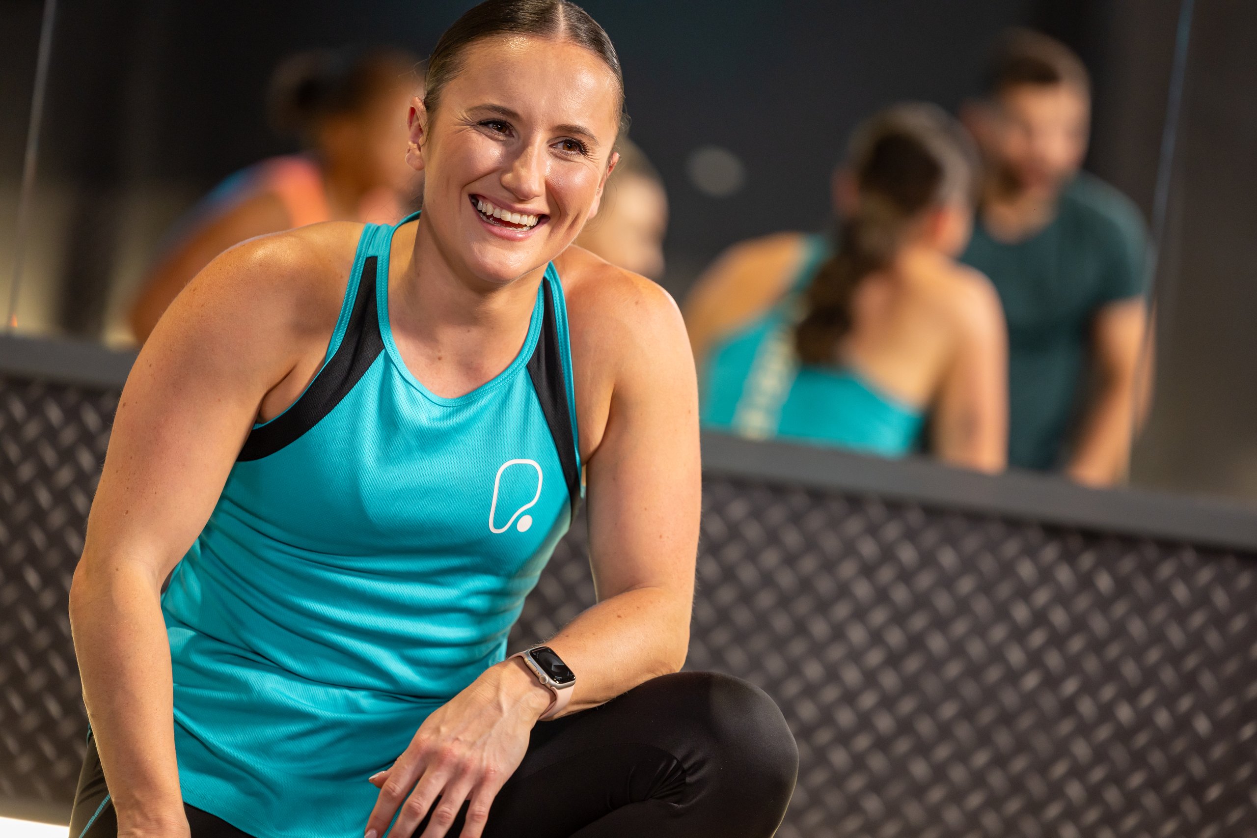 A smiling woman in a teal athletic top crouching in a gym with other people reflected in a mirror behind her.