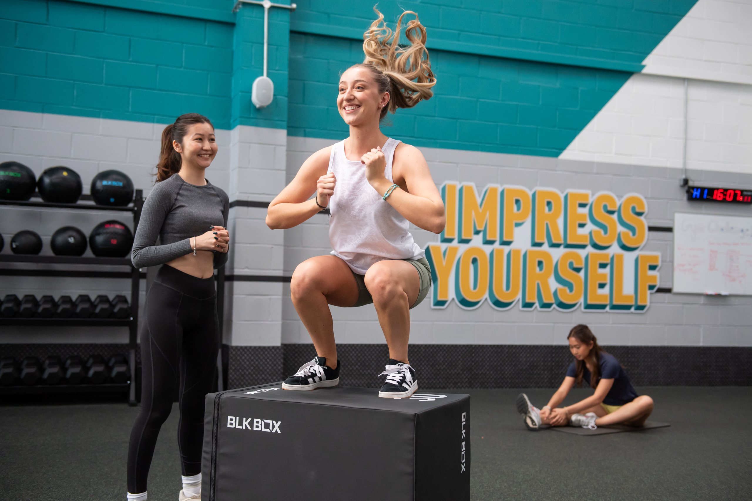 A woman jumps onto a gym box while another woman smiles and watches her closely.