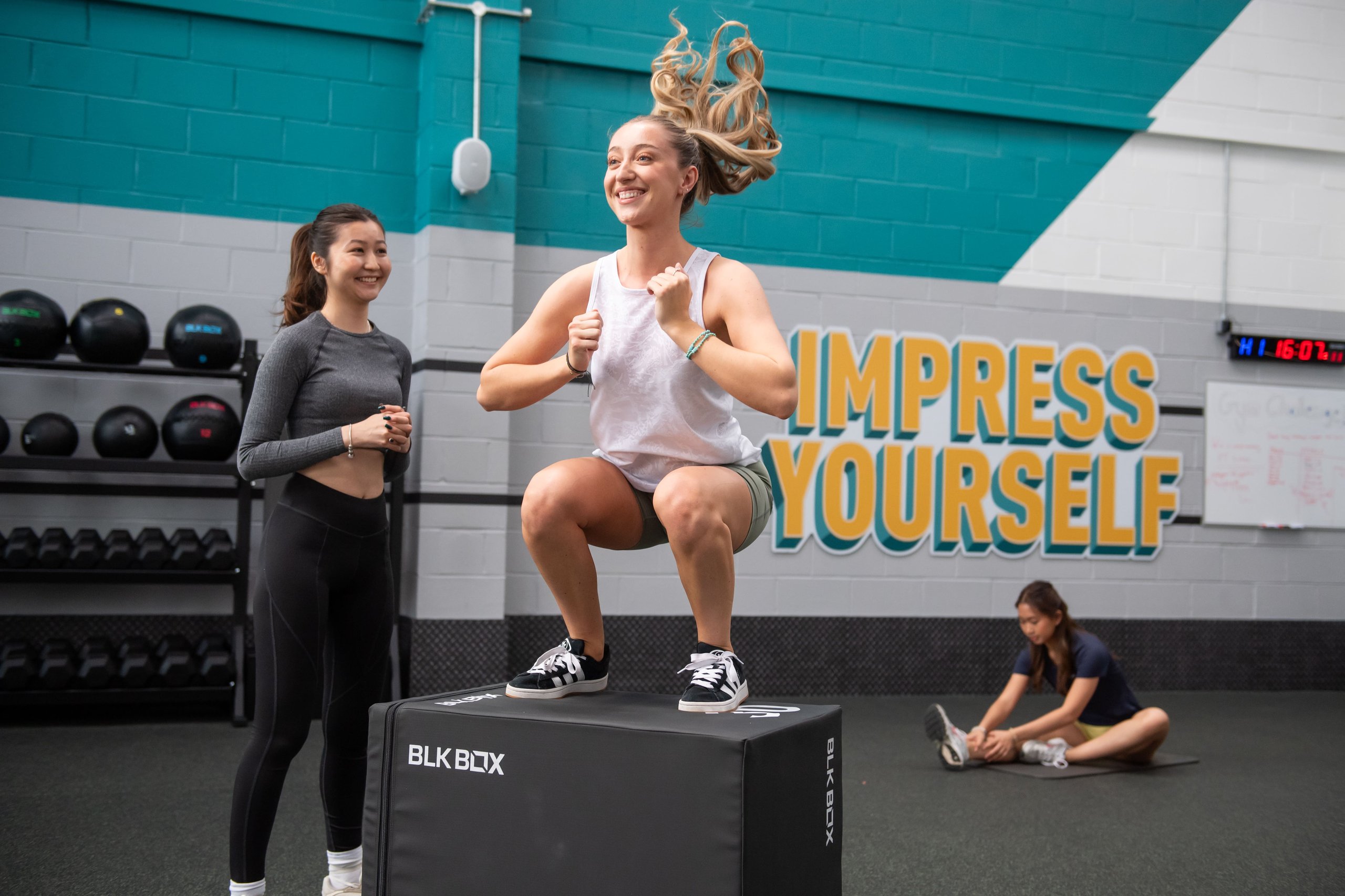 A woman jumps onto a gym box while another woman smiles and watches her closely.