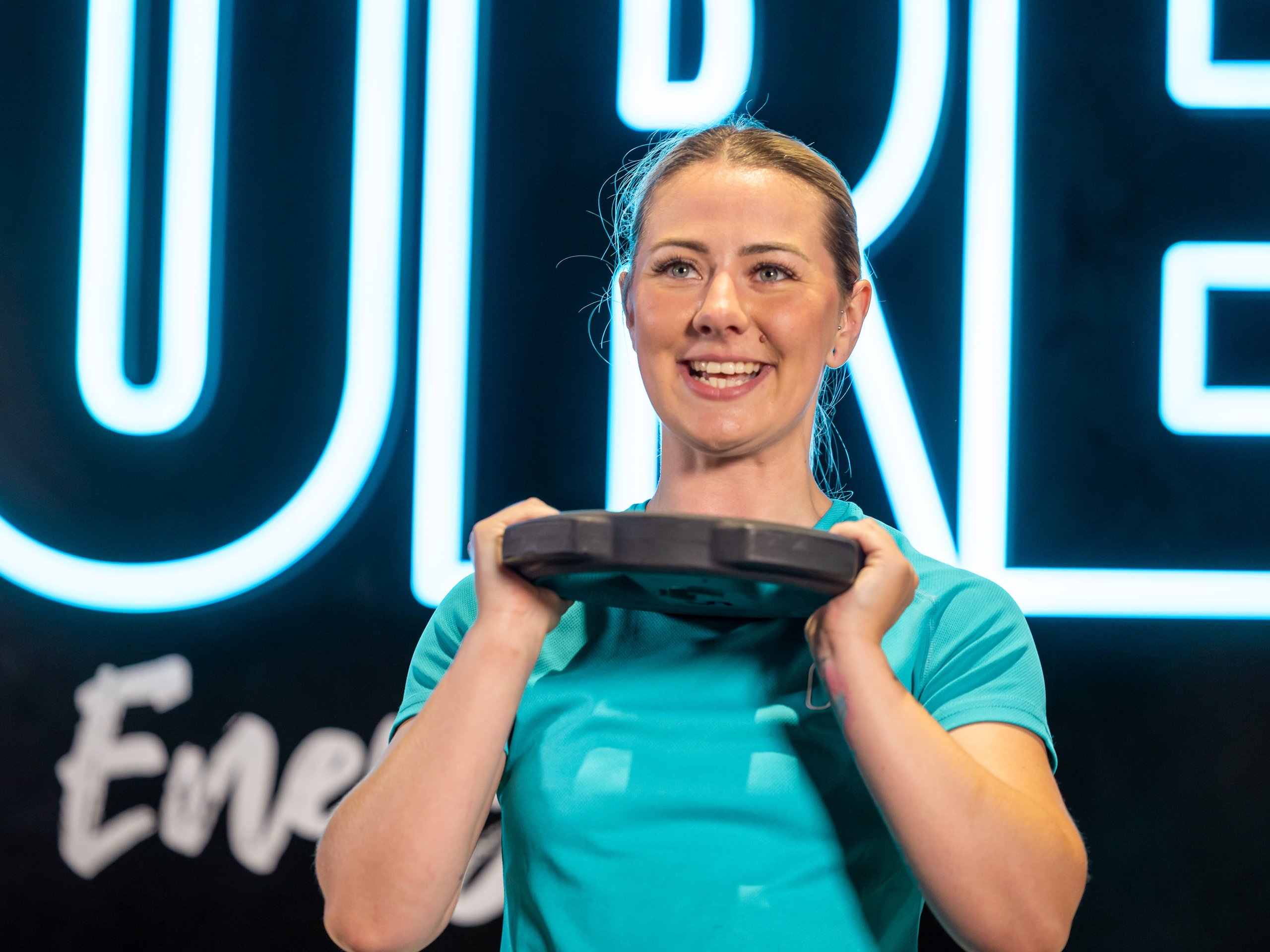 A smiling woman in a teal gym shirt lifting a weight plate in front of a glowing neon sign.