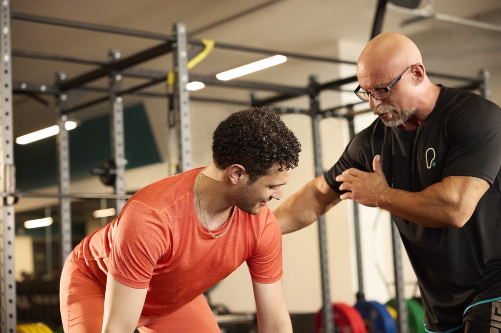 A gym instructor guiding a young man in an orange shirt with his exercise form.