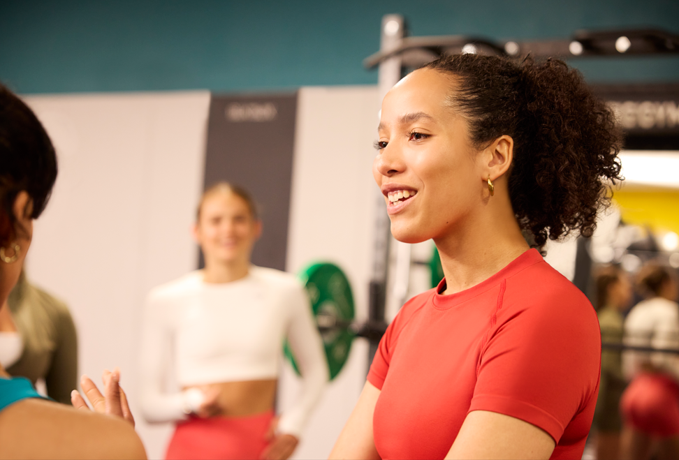 A smiling woman in a red shirt talking to friends inside a gym.